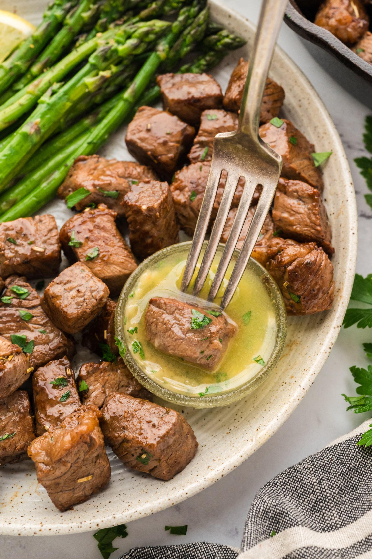 Steak Bites WEB-30 Steak bite being dipped in garlic butter sauce on plate next to asparagus.