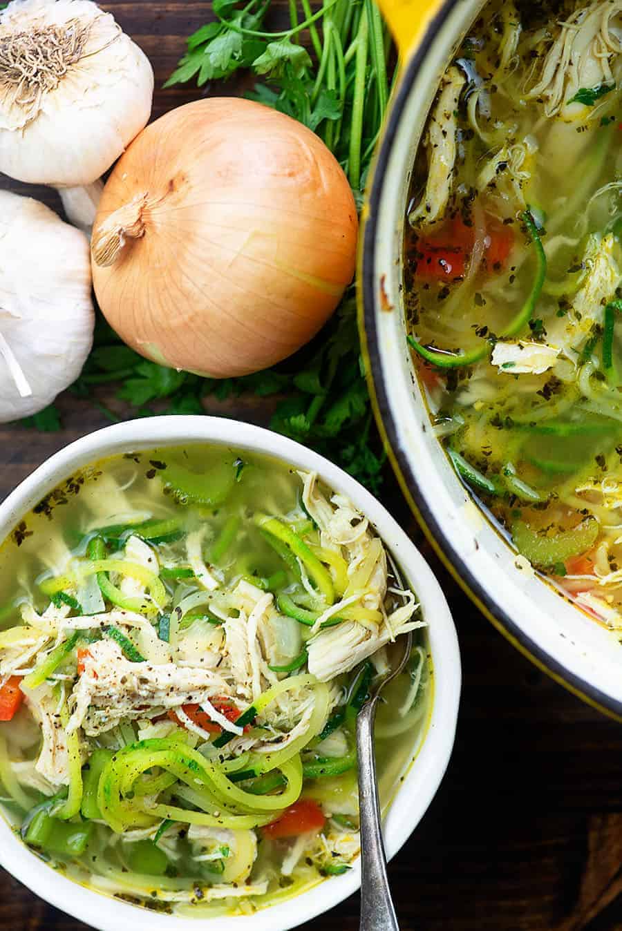 garlic and onions on a table next to a bowl of chicken soup and the pot it was served from