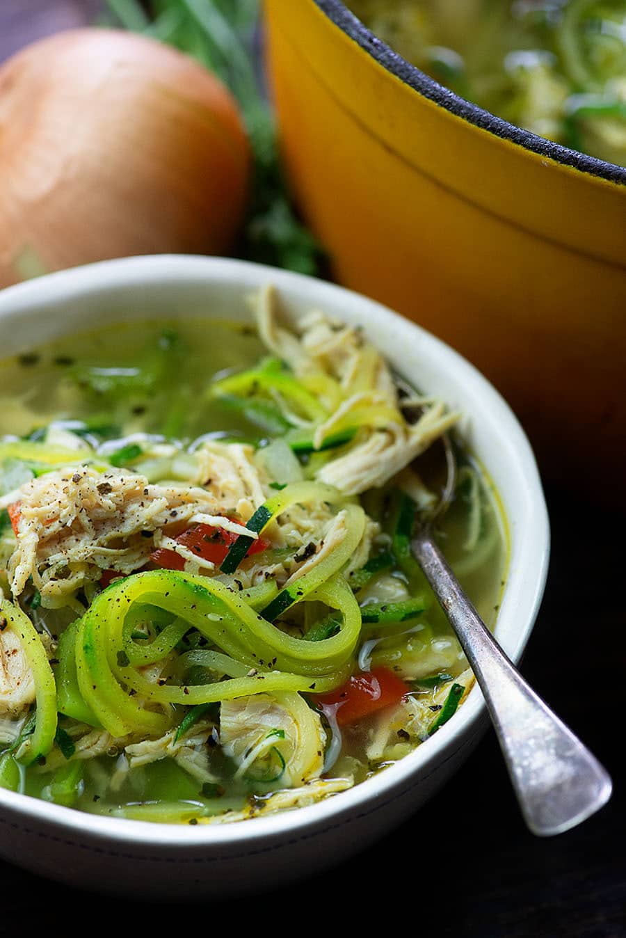 white bowl full of chicken zoodle soup with onion in background