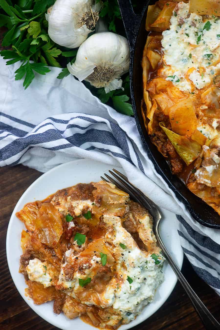 Lasagna on a white plate with fork.  skillet and ingredients above the plate