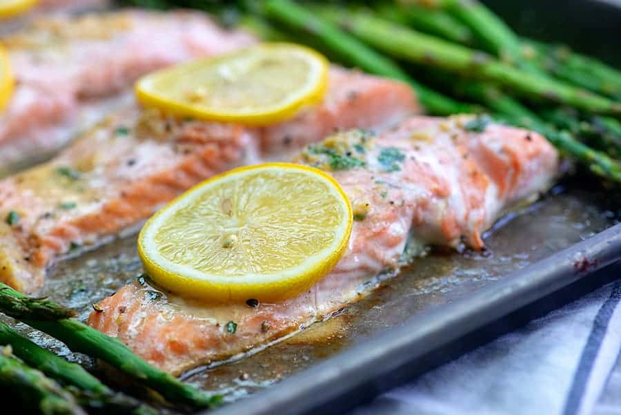 A close up of garlic butter salmon on a baking sheet in front of a pile of asparagus.