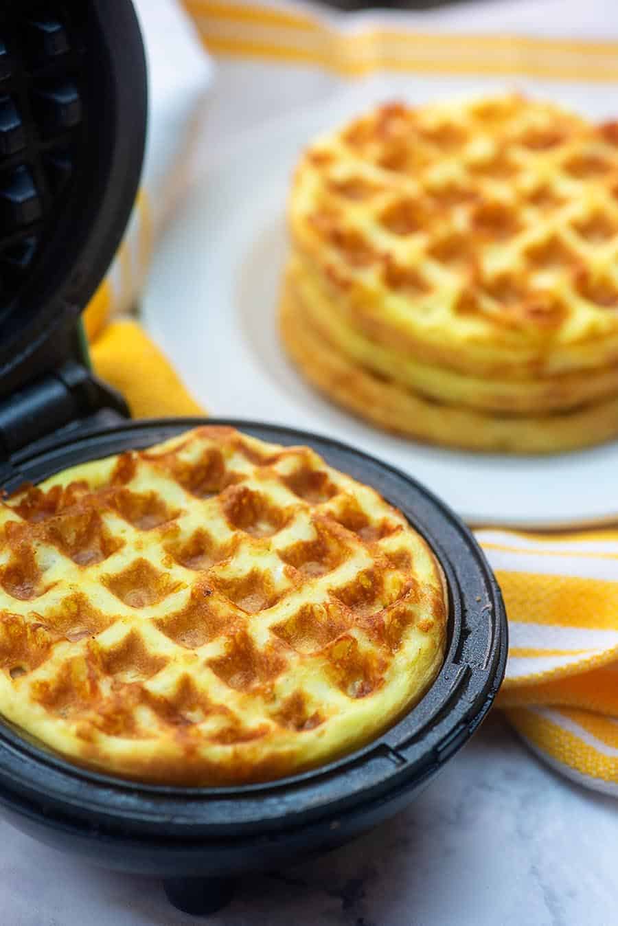 A chaffle in a mini waffle iron in front of a plate of stacked chaffles