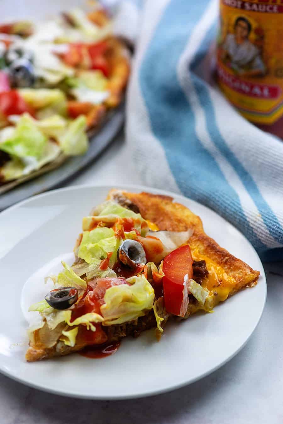 white plate with a piece of taco pizza on it and hot sauce in the background