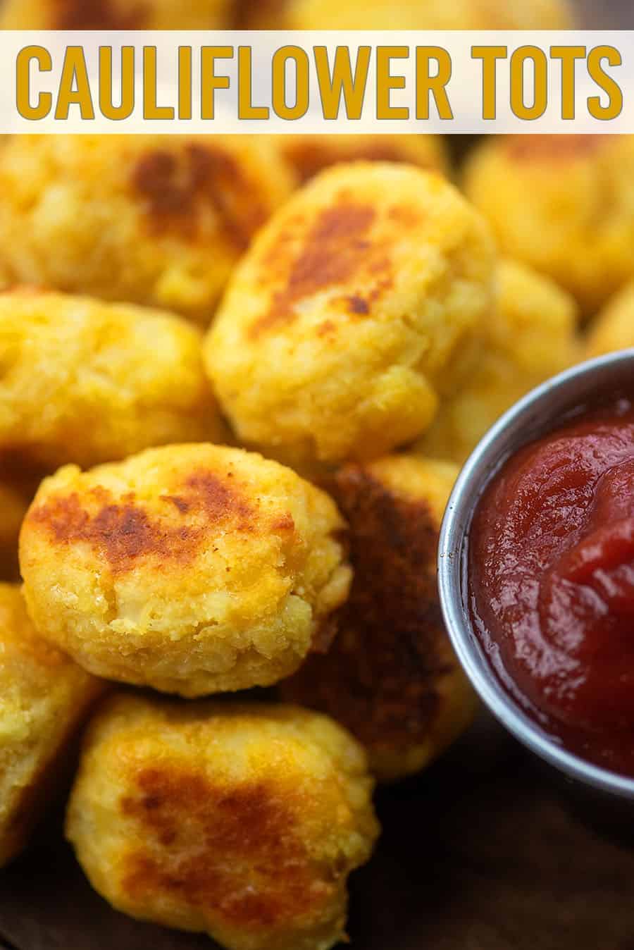 A close up of cauliflower tots piled up next to a cup of ketchup