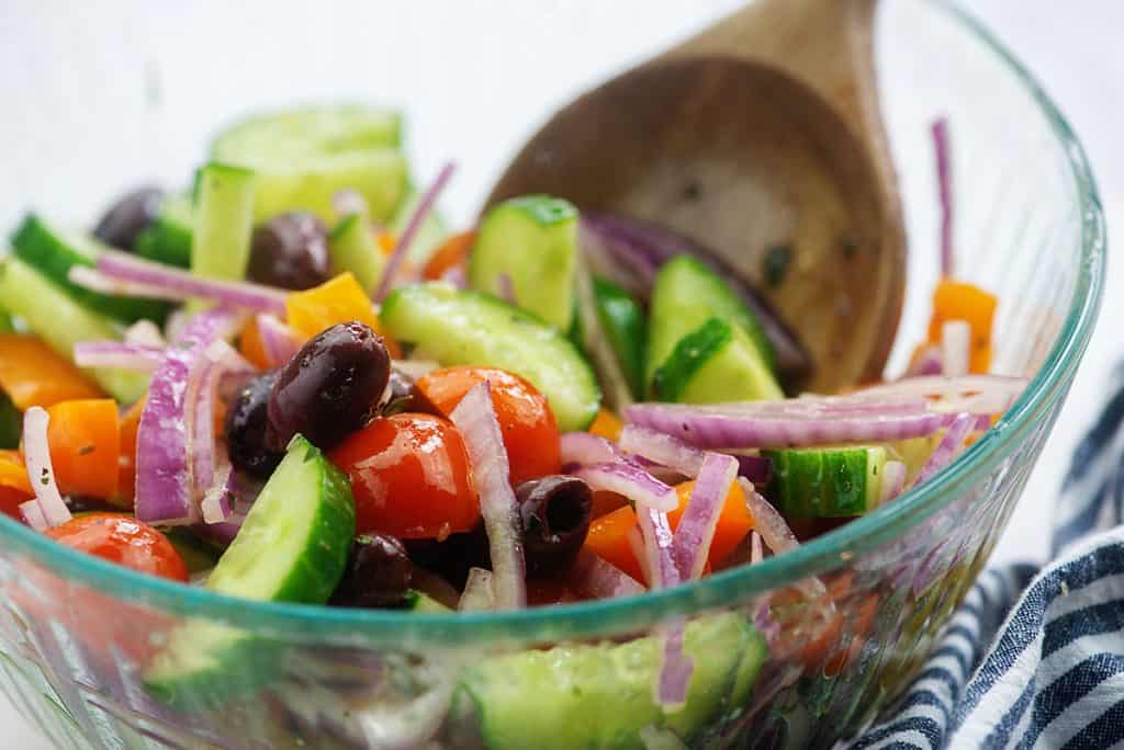 Greek cucumber salad in glass bowl with dressing and wooden spoon