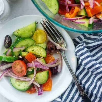 cucumber, tomato, red onion, and olives with Greek salad dressing on white plate with blue striped napkin