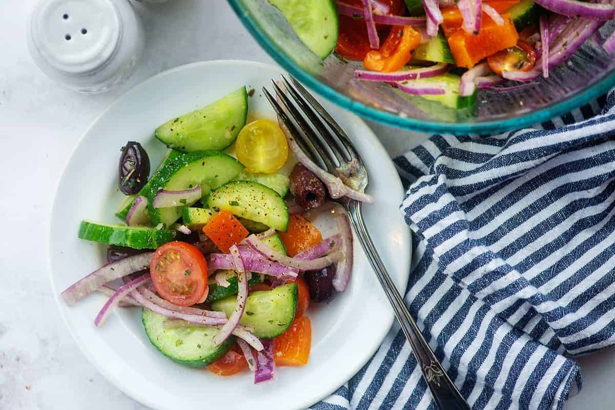 cucumber, tomato, red onion, and olives with Greek salad dressing on white plate with blue striped napkin