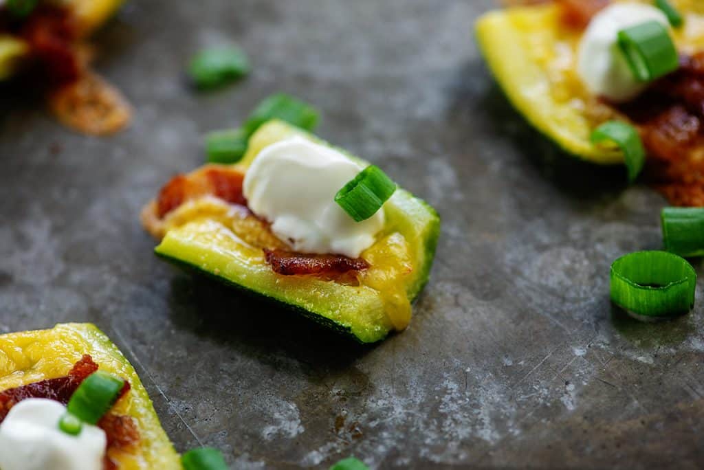 zucchini slices topped with cheddar, bacon, sour cream, and scallions on baking sheet