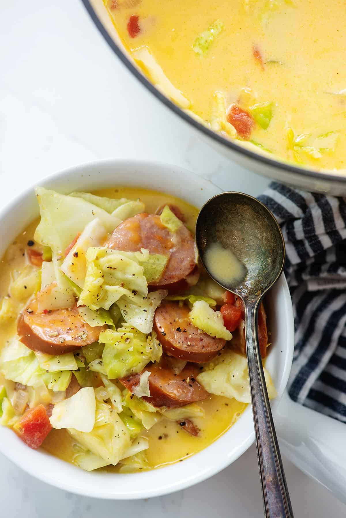 cabbage soup with sausage in white bowl next to pot of soup