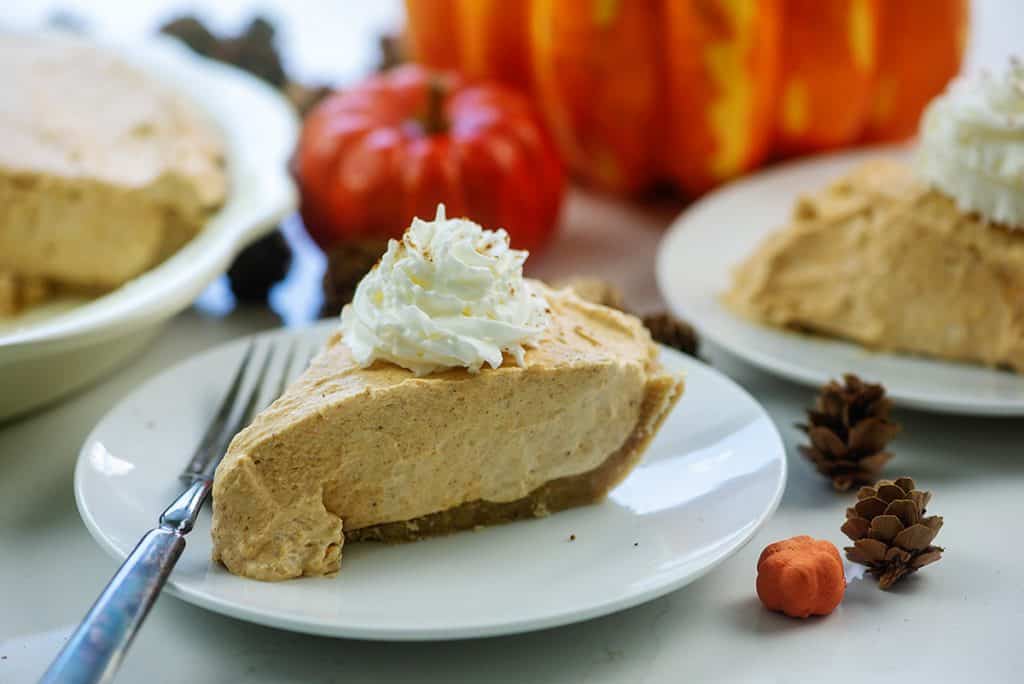 slices of keto pumpkin cream pie on white plates surrounded by pumpkins and pinecones