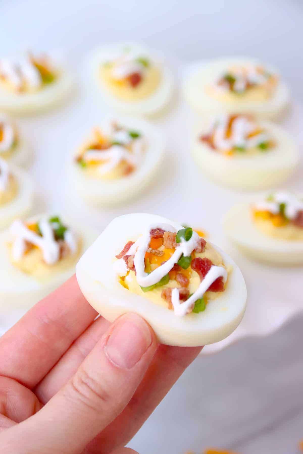a person holding a deviled egg topped with bacon and sour cream.