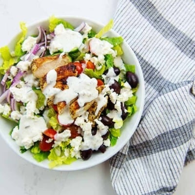 overhead view of Greek salad with chicken and tzatziki dressing in white bowl.
