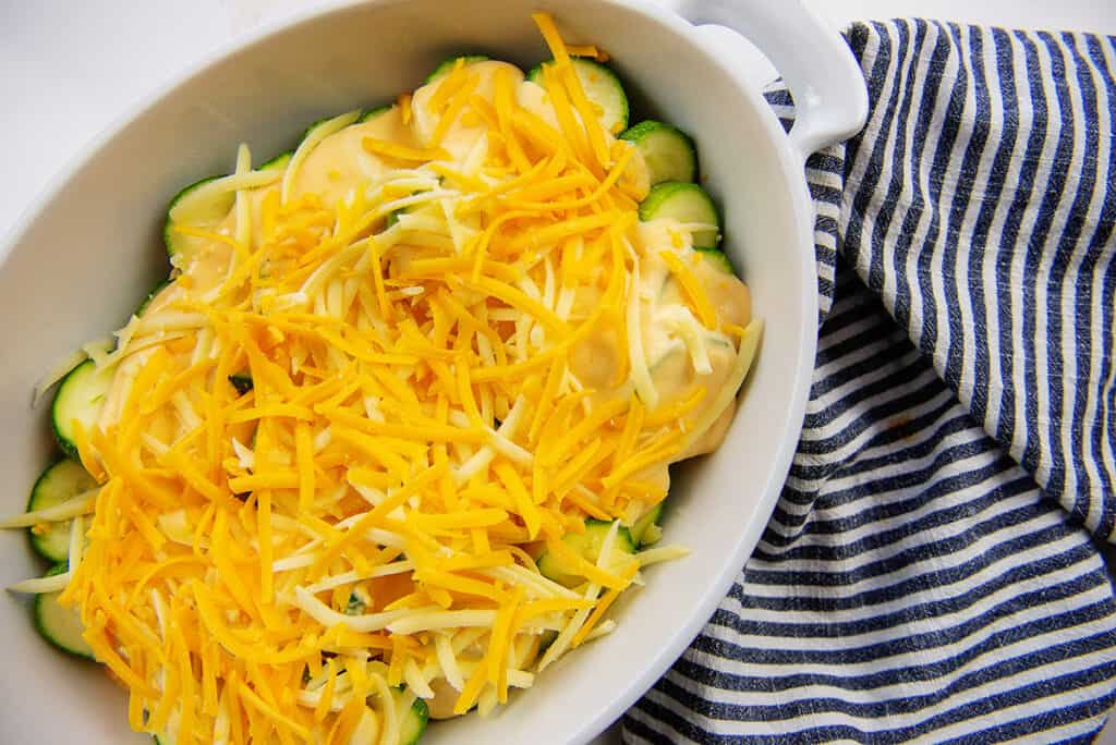 zucchini, cheese sauce, and shredded cheese layered together in white casserole dish.