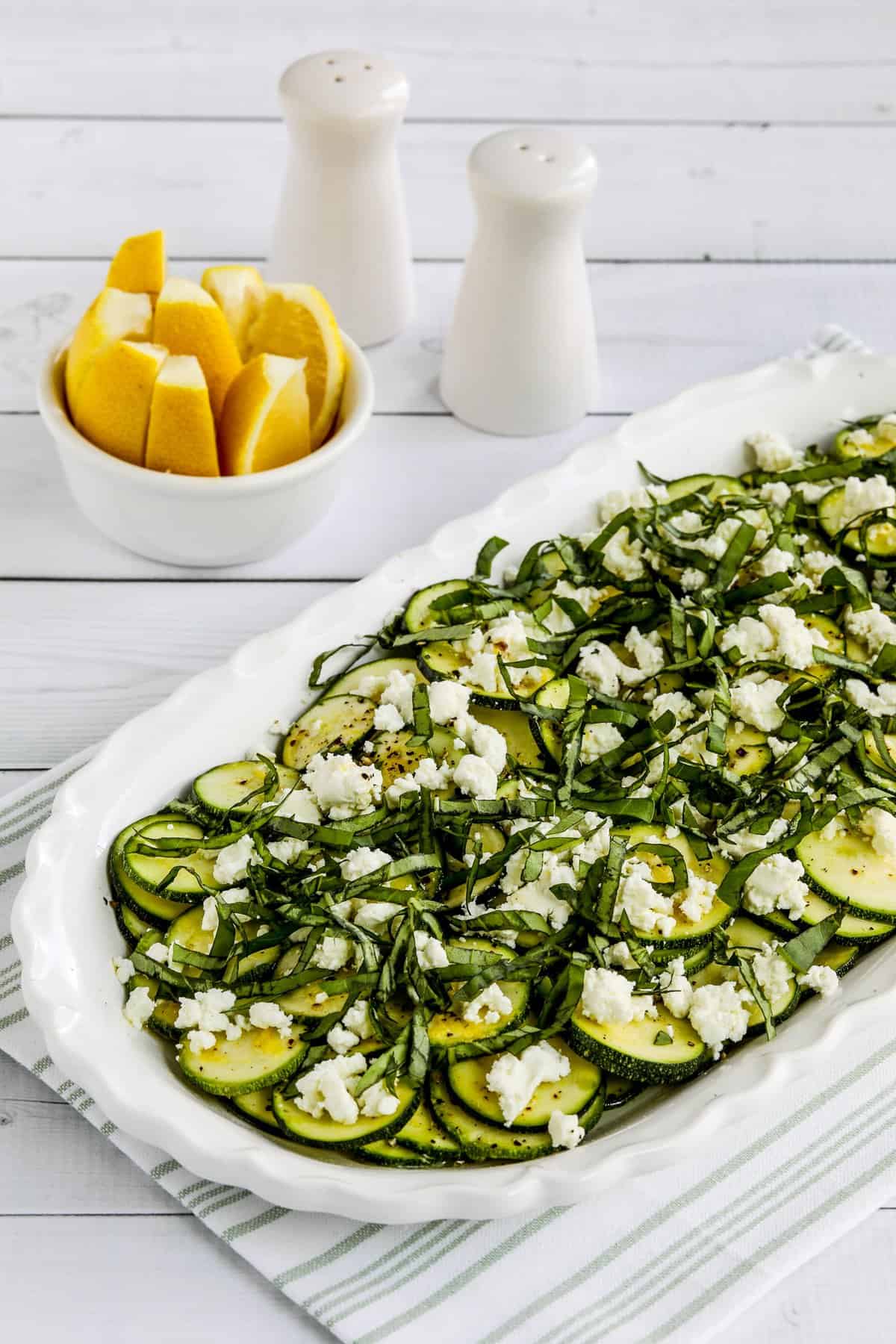 Zucchini carpaccio with feta cheese on a large white serving dish next to salt and pepper shakers and a bowl with fresh lemon wedges.