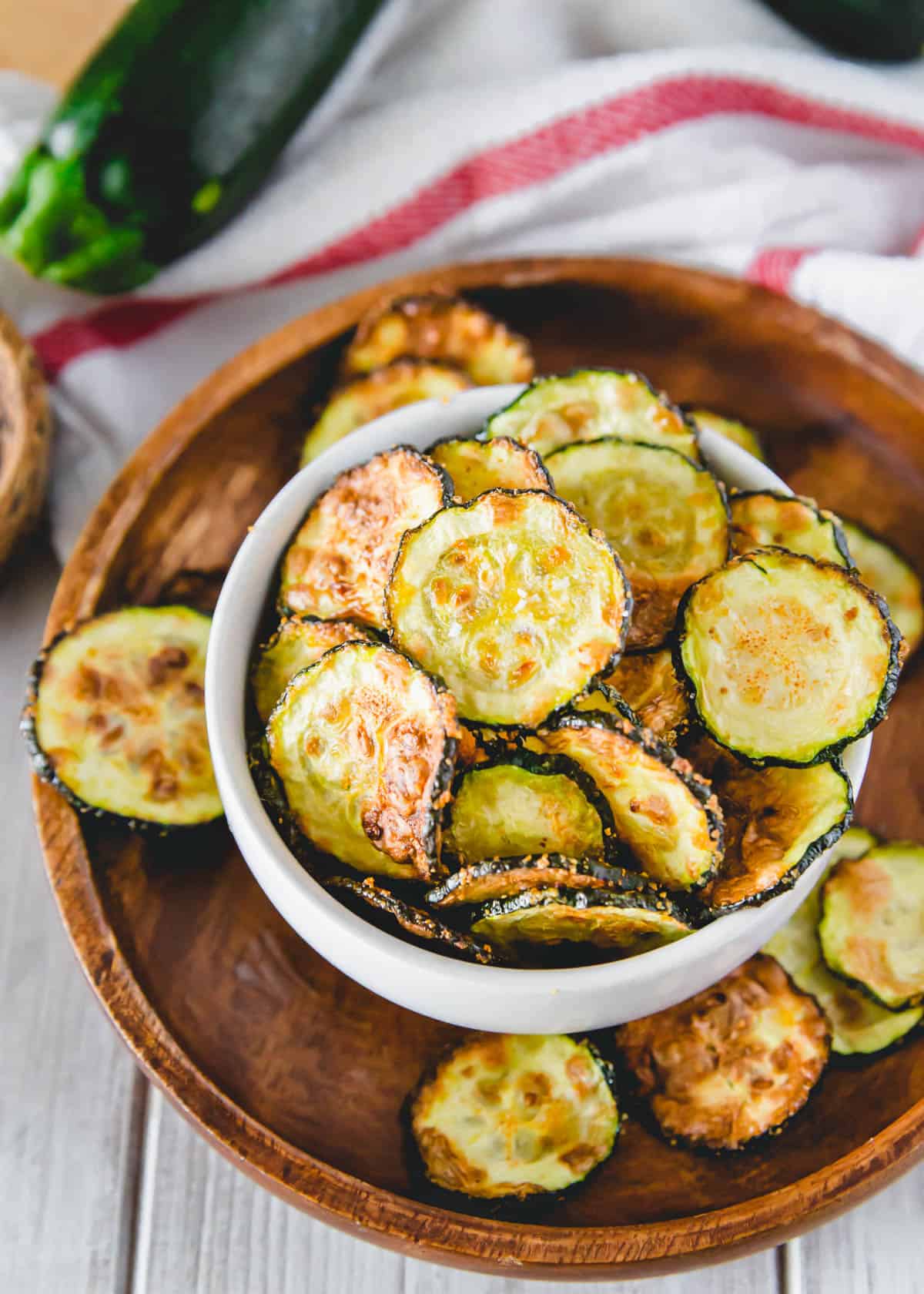 A white bowl on a wooden platter containing air fryer zucchini chips.