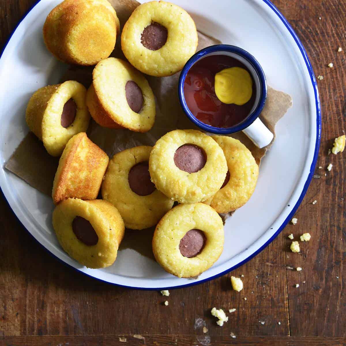 A top down view of low carb corn dog bites on a white and blue plate with a dish containing mustard and ketchup.