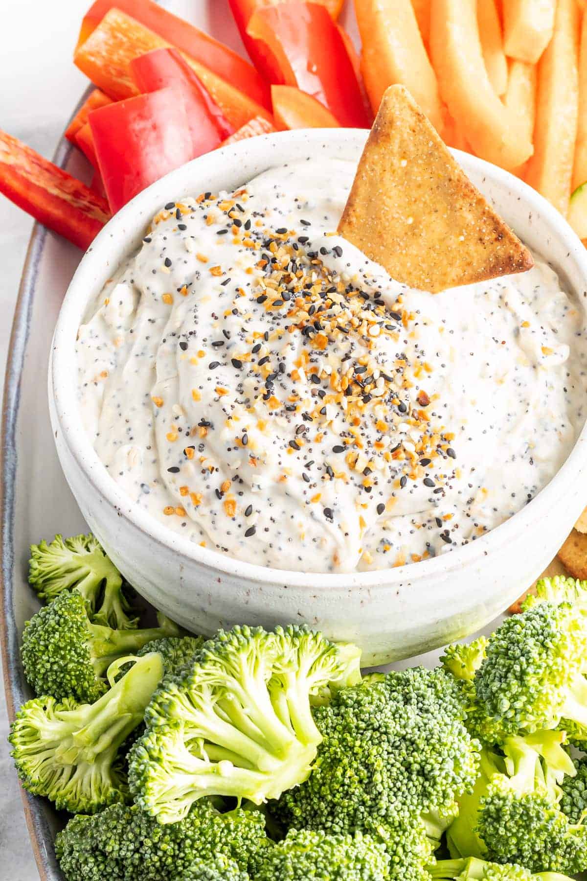 A white bowl containing everything bagel dip with pita chips, fresh broccoli, and bell peppers on a tray next to the bowl.