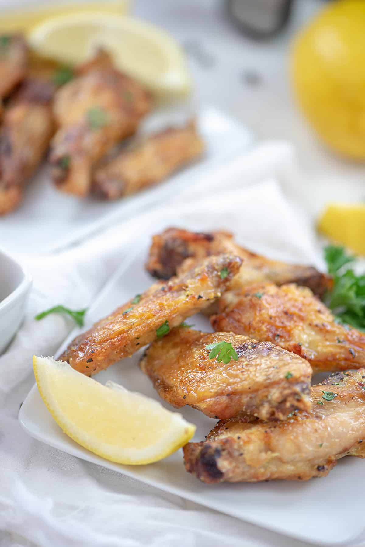 Lemon pepper chicken wings on a white plate next to a wedge of lemon with another plate in the background.