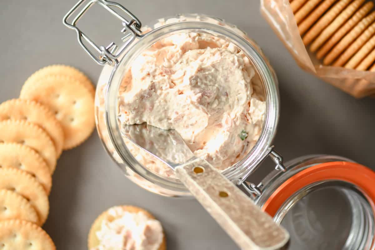 A top down view of smoked trout dip in a glass jar next to crackers on a table.