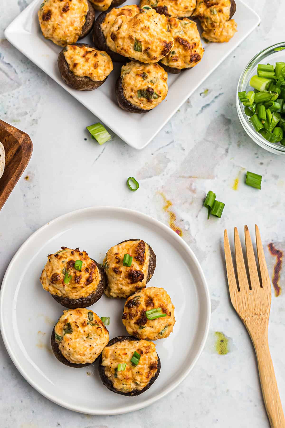 A top down view of plates containing keto stuffed mushrooms next to a wooden fork and a bowl containing chopped green onion.