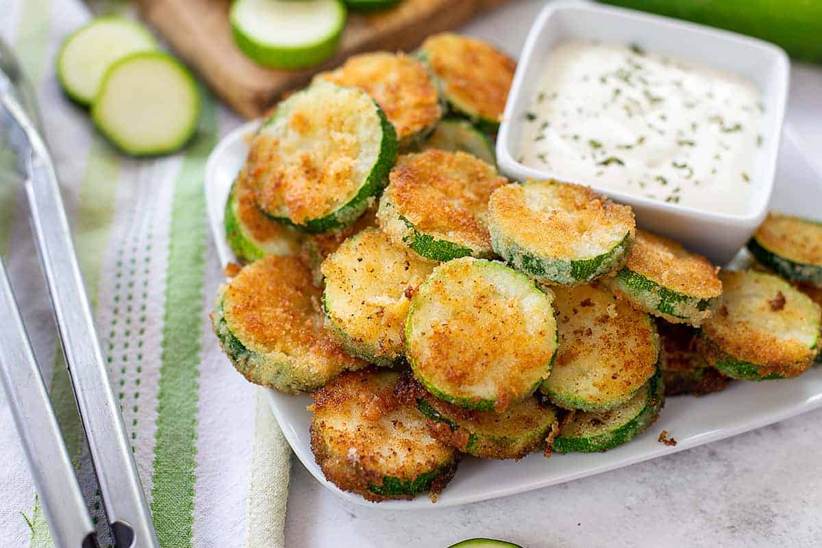 Fried zucchini rounds on a white plate next to ranch dipping sauce in a white bowl.