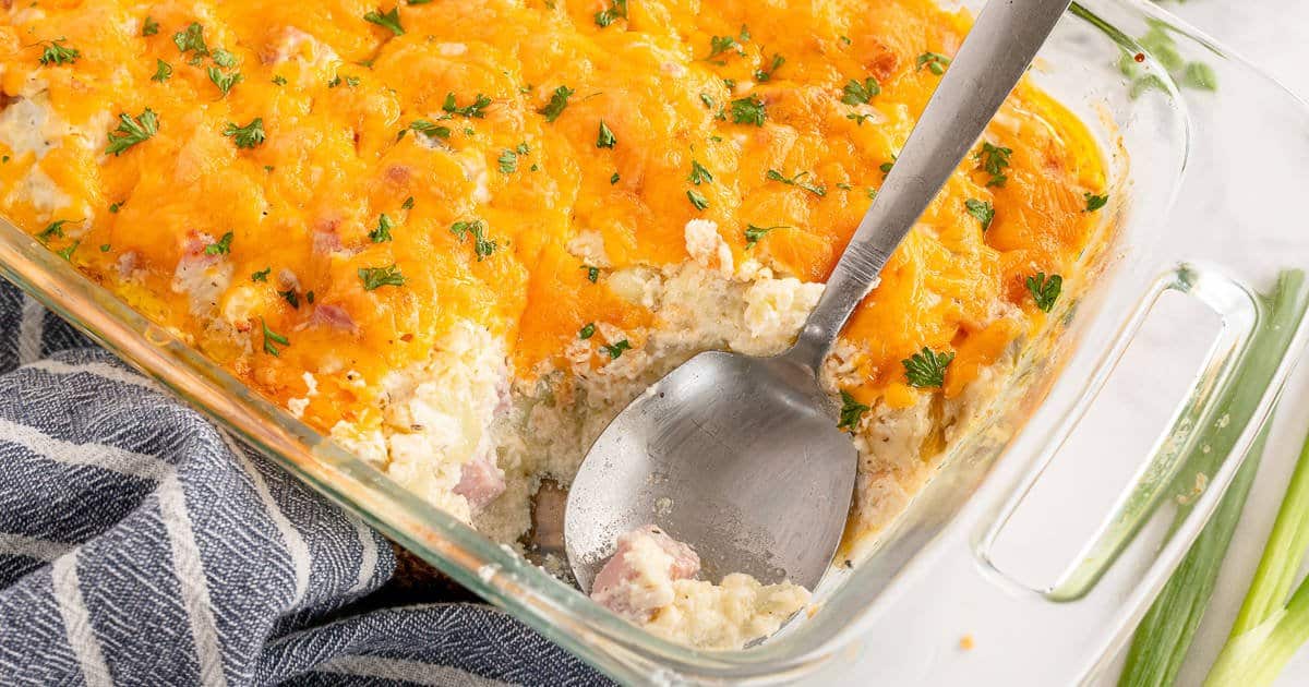 A glass baking dish containing cauliflower ham casserole with a large spoon scooping part of it out of the dish.