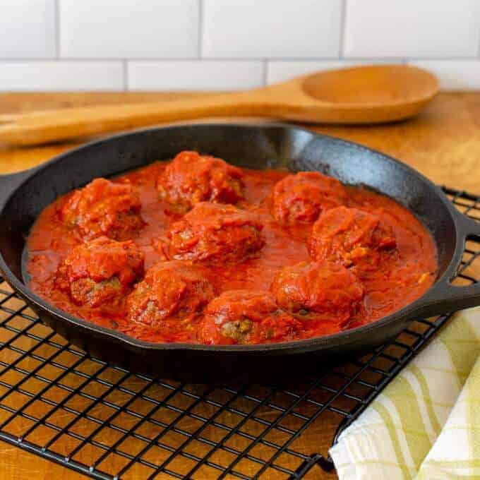A cast iron skillet containing meatballs without bread crumbs in marinara sauce on a cooling rack in front of a wooden spoon on a table.
