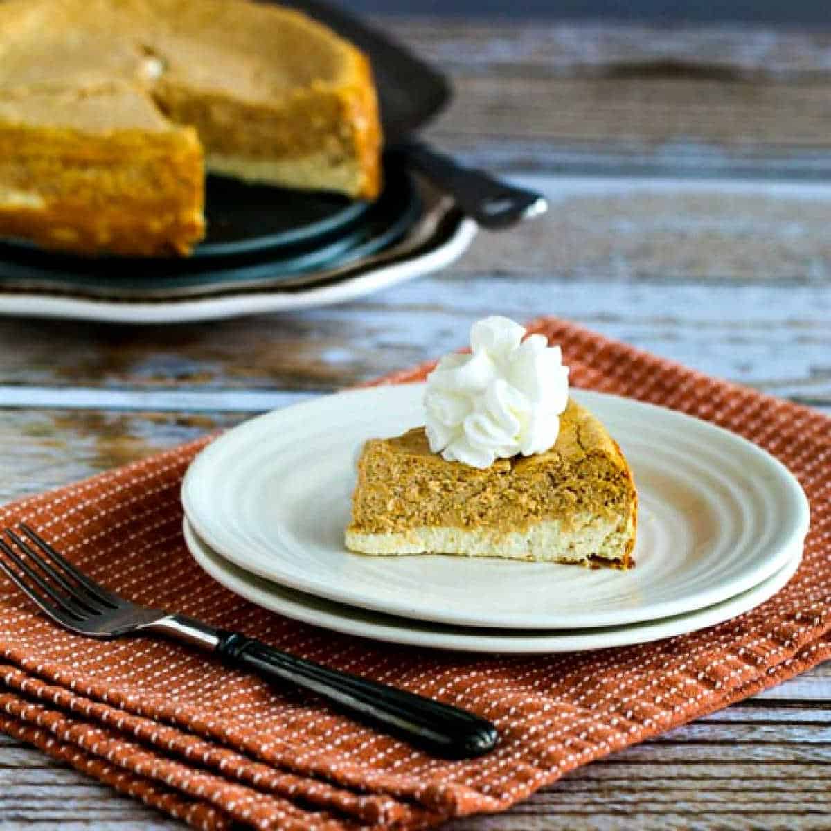 A slice of pumpkin cheesecake on a white plate with the rest of the cheesecake in the background.