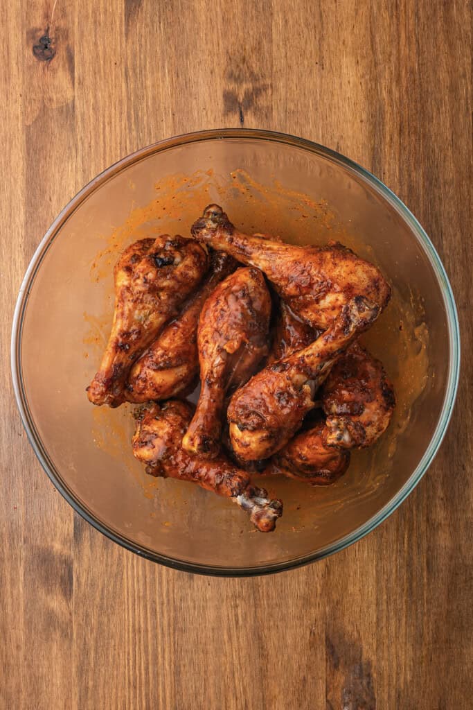 Baked chicken drumsticks tossed in buffalo sauce in glass mixing bowl.