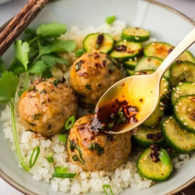 Sauce being drizzled over keto Asian turkey meatballs on plate with cauliflower rice and cucumber salad.