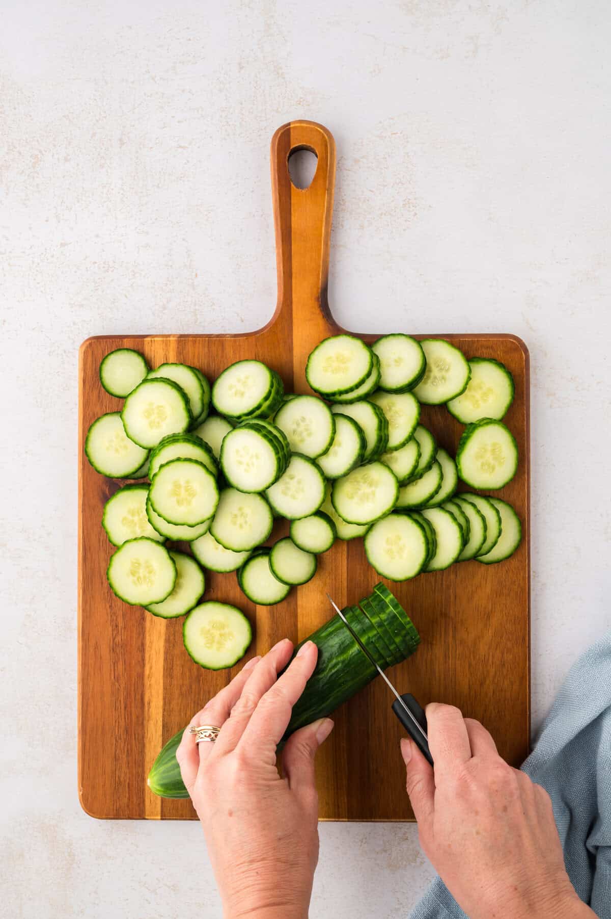 Cucumbers being thinly sliced on wooden cutting board.