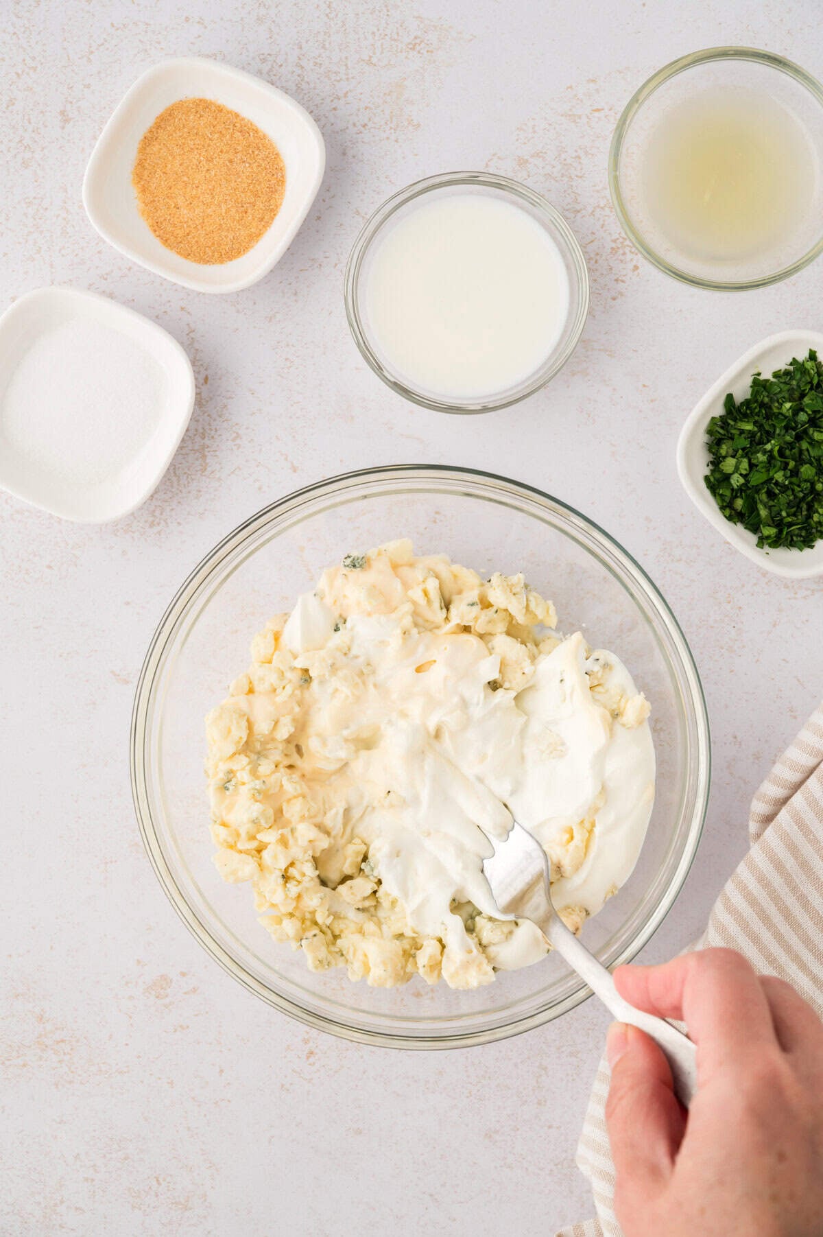 A hand using a fork to mash blue cheese into mayonnaise and sour cream.